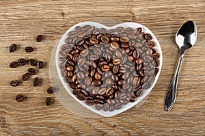 Scattered beans, roasted coffee beans in saucer in heart shape, spoon on wooden table. Top view