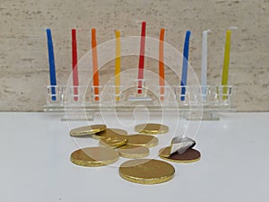 Scattered chocholate coins with a glass hannukiah filled with colourful candles in the background