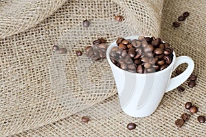 Scattered coffee beans and white exclusive porcelain cup filled with fragrant roasted coffee beans on jute sacking