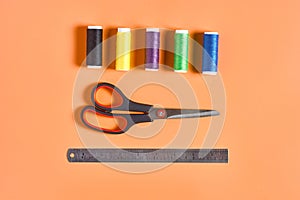 Scattered sewing accessories metal ruler, scissors and spools with colorful threads on orange tailors desk in workshop