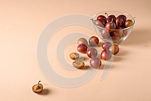 Scattered sliced half ripe sweet plum fruits with water drops near to plums in glass bowl on cream colored background