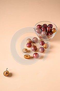 Scattered sliced half ripe sweet plum fruits with water drops near to plums in glass bowl on cream colored background