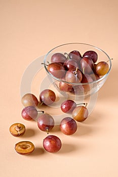 Scattered sliced half ripe sweet plum fruits with water drops near to plums in glass bowl on cream colored background