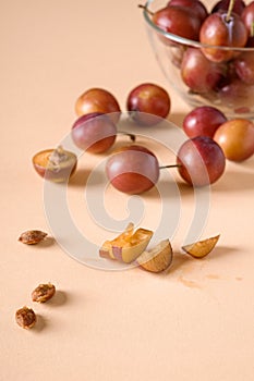 Scattered sliced half ripe sweet plum fruits with water drops near to plums in glass bowl, on cream colored background