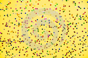 Scattered star shaped colorful glittering confetti over yellow background.