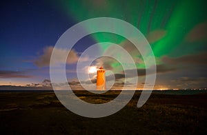 Scene of the night sky with northern lights and a moon behind the clouds over the white lighthouse