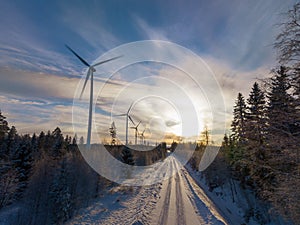 Scenic aerial photo over forest winter road with windmills standing in row at the left side in forest. Road in the middle of view