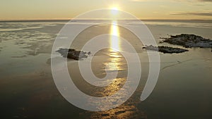 Scenic fly up from snow covered big stones at coastline to winter sea with thin ice floes illuminated by Sunset. Sunlight path on