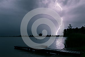 Scenic night view of a pier at the shore of a lake while lightning strikes in the background