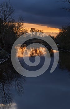 Scenic sunset sky with dark clouds and leafless trees reflecting in water