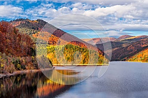 Autumn colored forest reflecting to alpine lake in Transylvanian Alps during warm October light against dramatic sky