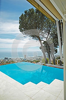 Scenic view of infinity swimming pool overlooking city and ocean in background. Luxury outdoor blue pool on a tiled deck