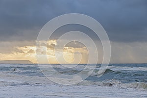 A scenic view of a raging sea with huge crashing waves with a majestc ray of light coming through a cloudy stormy sky with some