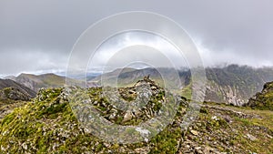 A scenic view of a rocky mountain summit with a stony cairn and mountain range in the background under a stormy grey cloudy sky