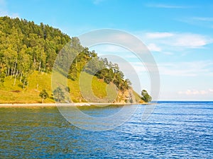 Scenic view of summer calm lake Baikal on the edge of the forest and green hill, against the blue sky