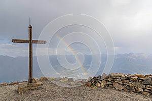 A scenics view of a mountain summit cross with a majestic double rainbow in the background under a stormy and rainy weather