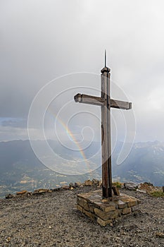 A scenics view of a mountain summit cross with a majestic rainbow in the background under a stormy and rainy weather conditions