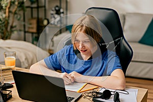 School age boy with long hair spends time in room at desk on chair doing homework in front of computer, remote teaching