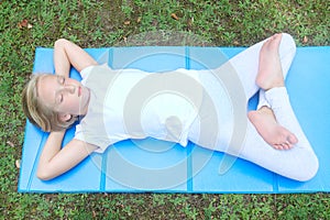 School-age girl in light clothes has a rest and plays sports, her eyes closed, she lies on the mat in the park.