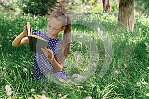 A school-age girl reading a book sitting on the grass in the park. Front view.