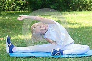 School-age kid girl in light clothes take sport exercise on a mat in the park. Outdoors workout.