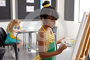 In school, during art class, a young biracial girl with dark hair is holding a paint palette, painti