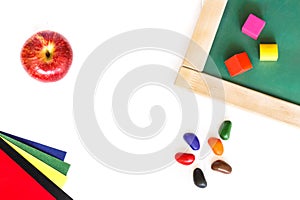School board, colored blocks, red apple, wax crayons, colored paper lying on a white wooden background.