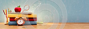 School books and school accessory on wooden table in front of empty blue chalkboard with copy space.