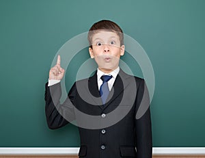 School boy in black suit show finger up gesture and wonder, point on green chalkboard background, education concept