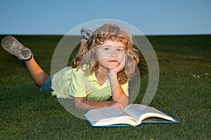 School boy with a book having a rest outdoor. Clever child boy reading book laying on grass on grass and sky background