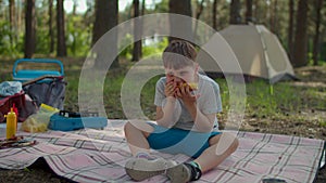 School boy eating grilled corn holding in hands during summer family camping vacation with tent in forest. Slow motion