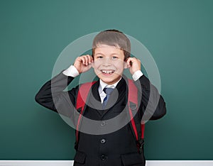 School boy make faces in black suit on green chalkboard background with red backpack, education concept