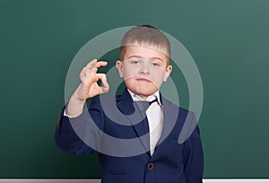 School boy show ok sign, portrait near green blank chalkboard background, dressed in classic black suit, one pupil, education conc