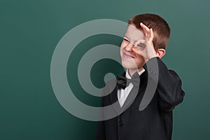 School boy show ok sign, portrait near green blank chalkboard background, dressed in classic black suit, one pupil, education conc