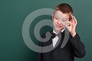 School boy show ok sign, portrait near green blank chalkboard background, dressed in classic black suit, one pupil, education conc