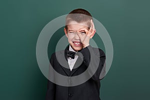 School boy show ok sign, portrait near green blank chalkboard background, dressed in classic black suit, one pupil, education conc