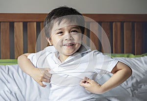 School boy sitting in bed and try to wearing his cloth with smiling face,Cute kid boy getting dressed and get ready for school,