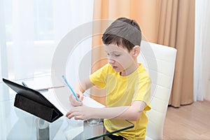 School boy in yellow t-shirt sitting at the table with digital tablet studying at home. Distance learning online