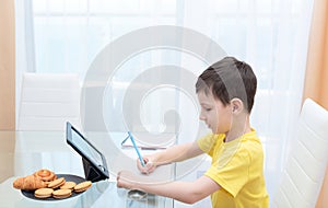 School boy in yellow t-shirt sitting at the table with digital tablet studying at home. Distance learning online