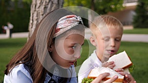 School children sitting in the park on the grass and holding large sandwiches.