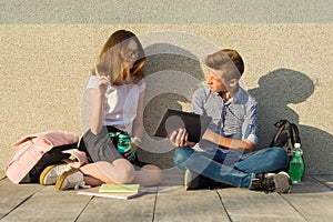 School children teens sit on the gray wall, read textbooks, drink water, look at the tablet.The girl is looking at the tablet and