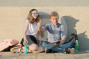 School children teens sit on gray wall, read textbooks, drink water, look at textbook. Boy looking at book and pointing forefinger