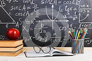 School desk in classroom, with books on background of chalk board with written formulas. Soncept Teacher`s Day