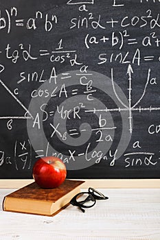 School desk in classroom, with books on background of chalk board with written formulas. Soncept Teacher`s Day