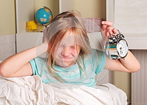 School girl hates waking up early in the morning. Blond girl holding alarm clock , yawning in bed covers ear with hand