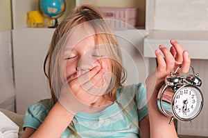 School girl hates waking up early in the morning. Blond girl holding alarm clock , yawning in bed covers ear with hand