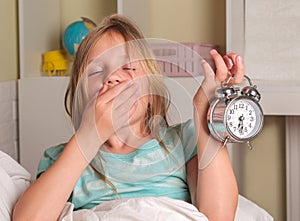 School girl hates waking up early in the morning. Blond girl holding alarm clock , yawning in bed covers ear with hand