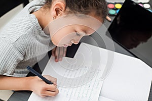 School girl writing on the notebook. Child follow lessons on the tablet