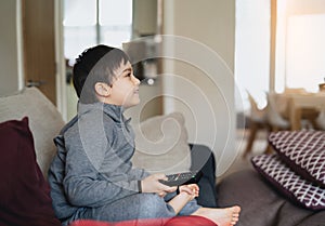 School Kid sitting alone on couch holding remote control and watching TV, Happy boy siting on sofa watching cartoon, Child