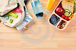 School lunch box with healthy food and school backpack with stationery on wooden background. Back to school concept. Top view.